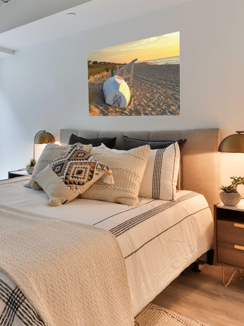 Bedroom with a bed featuring patterned pillows and a canvas print of an overturned Jersey Shore Beach Patrol boat and lifeguard stand on the sand at sunrise with beach grass and rolling dunes.