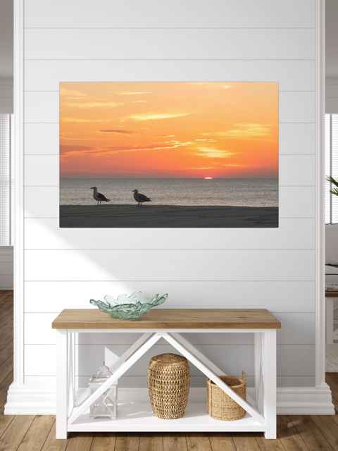 Canvas print of two seagulls standing on a sandy Jersey Shore beach during a vibrant orange sunrise over the Atlantic Ocean hanging on a white wall above a wooden bench.