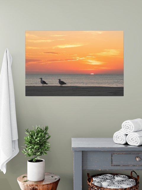 Canvas print of two seagulls standing on a sandy Jersey Shore beach during a vibrant orange sunrise over the Atlantic Ocean, hanging on a wall above a side table with towels and a plant.