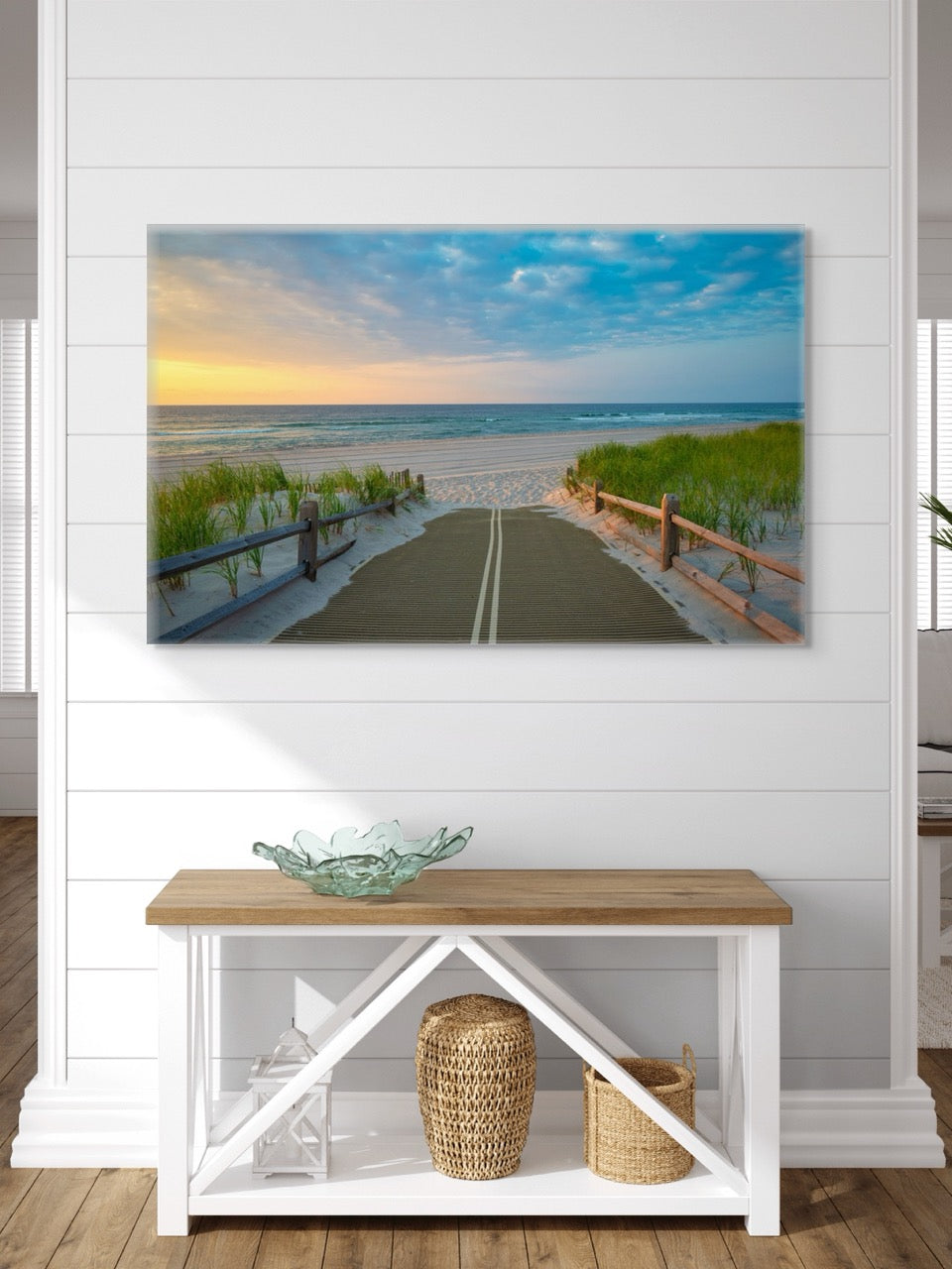 A canvas print of a beach path on the Jersey Shore shortly after sunrise over the ocean above a wooden bench with a glass bowl and baskets on a white wall.
