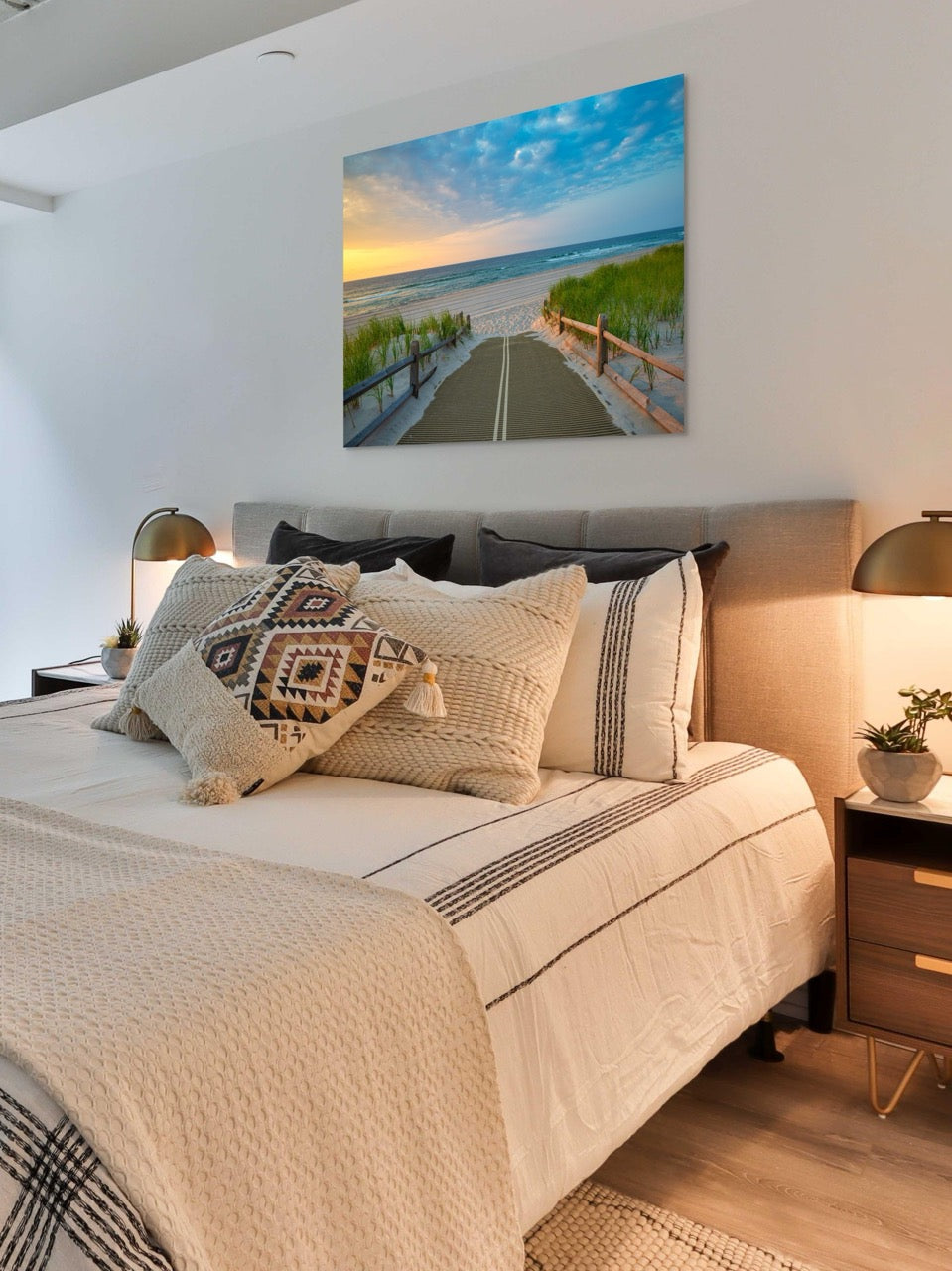 Neatly made bed with decorative pillows in a bedroom setting, featuring a large canvas print of a beach path on the Jersey Shore shortly after sunrise over the ocean