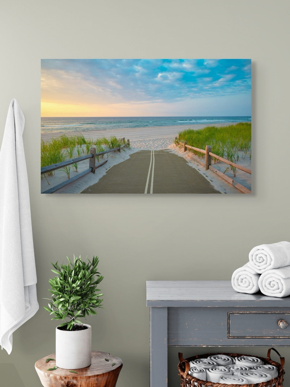 A canvas print of a beach path on the Jersey Shore shortly after sunrise over the ocean on a wall above a table with a plant and folded towels.