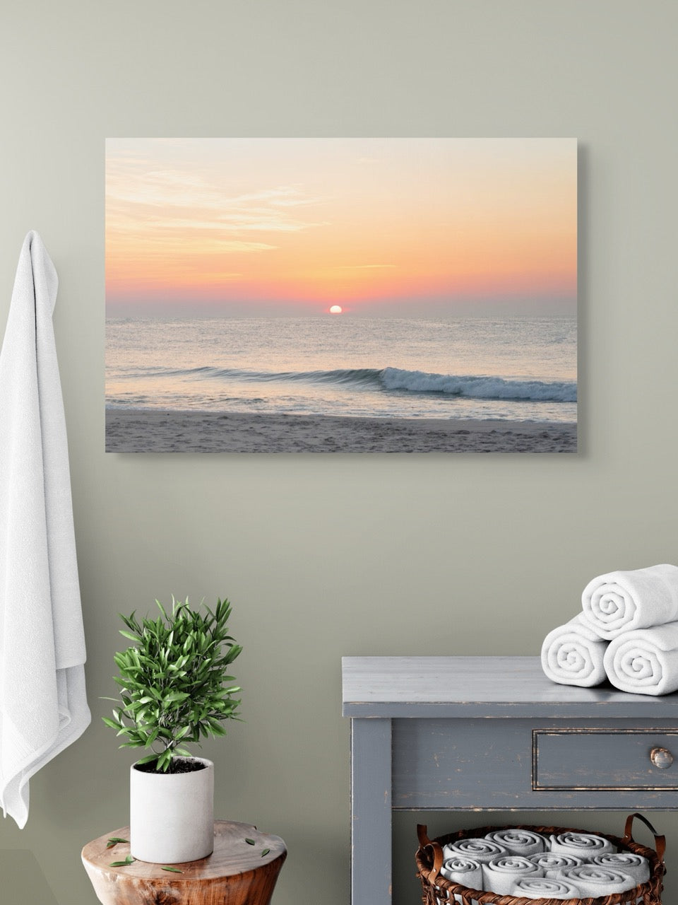 Large canvas print featuring a summer sunrise over the Atlantic Ocean as seen from a quiet beach on the Jersey Shore hanging on a wall above a small table with a plant and folded towels.