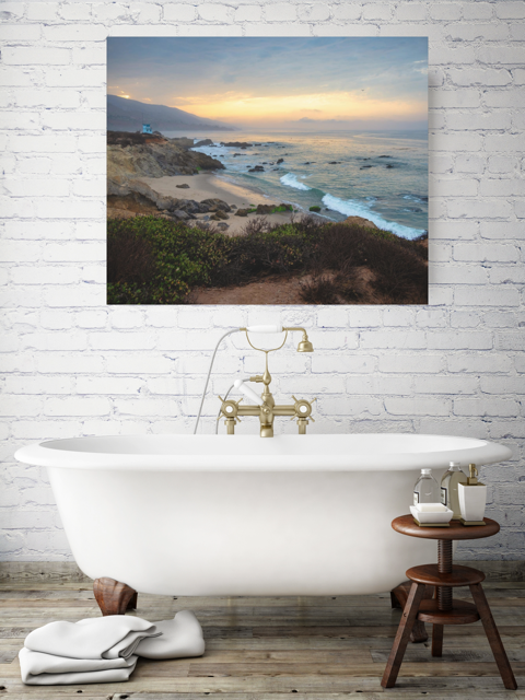 Bathroom with a freestanding bathtub, wooden stool with toiletries, and a large canvas print of the sunrise over the mountains as seen from the cliffs of Leo Carrillo State Beach near Malibu, California on the wall.