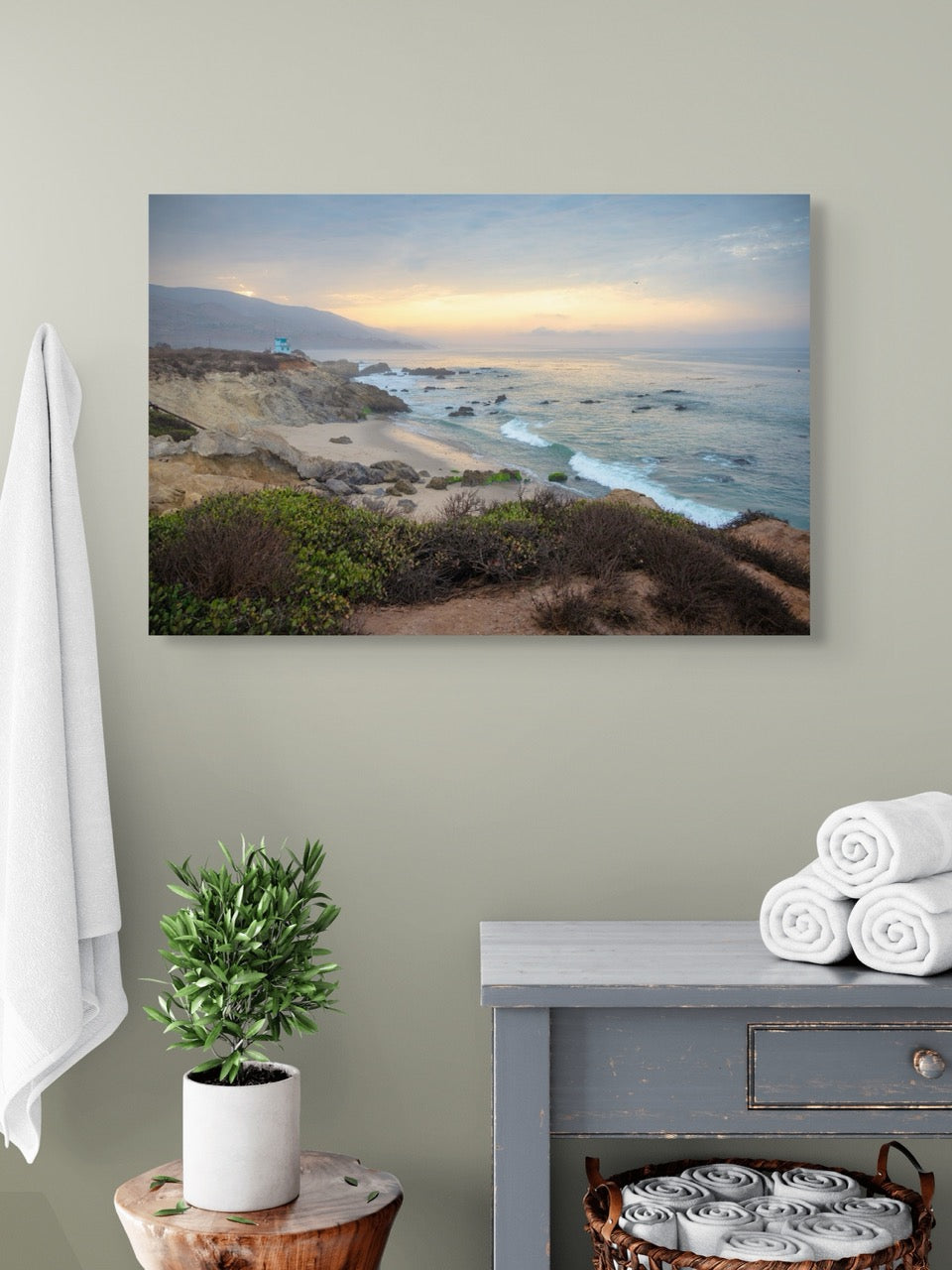 Canvas print of the sunrise over the mountains as seen from the cliffs of Leo Carrillo State Beach near Malibu, California hanging on a wall above a table with a plant and folded towels.