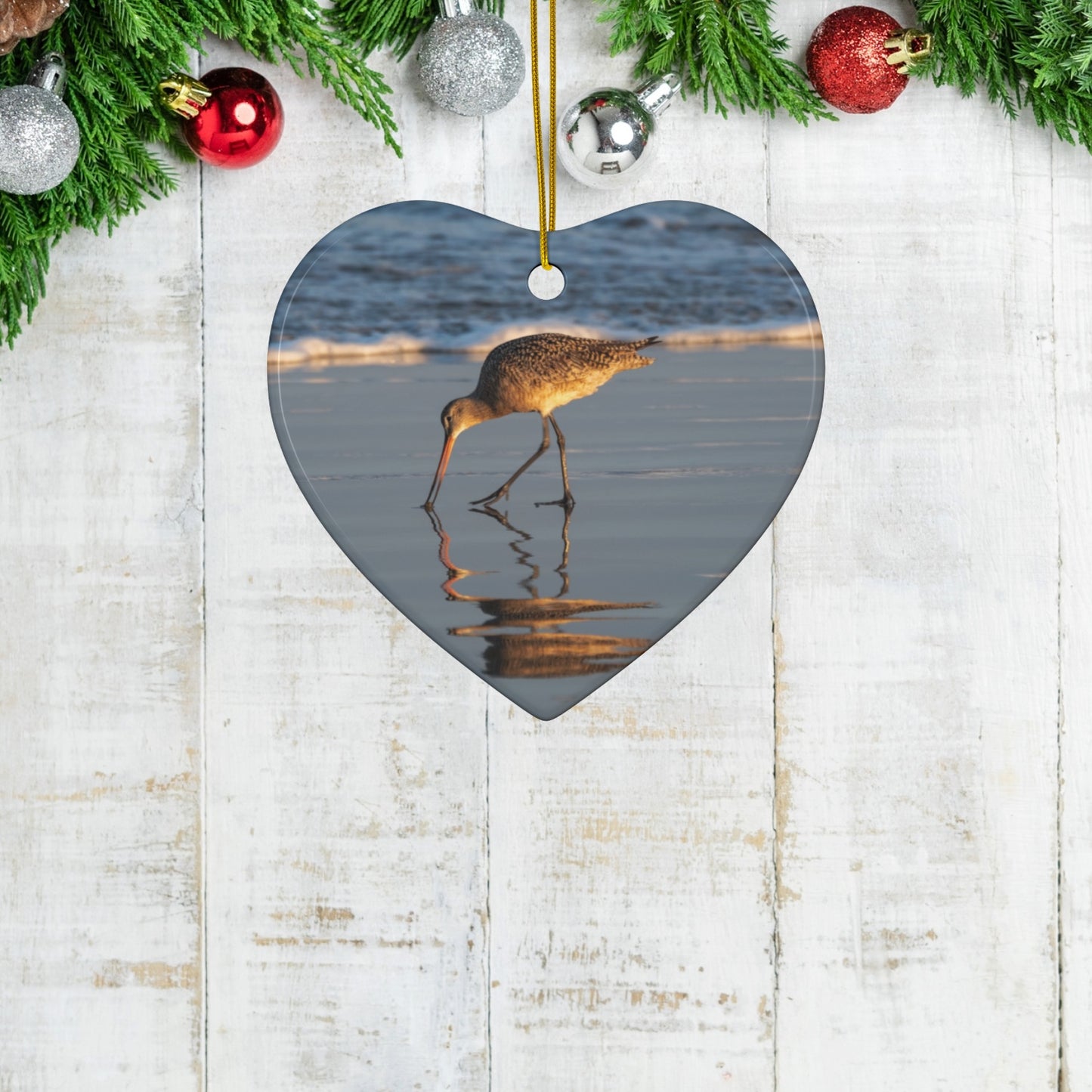 Heart-shaped ceramic holiday ornament featuring a photograph of a Long-billed Curlew mirrored in the wet sand of a shoreline under a soft golden sun
