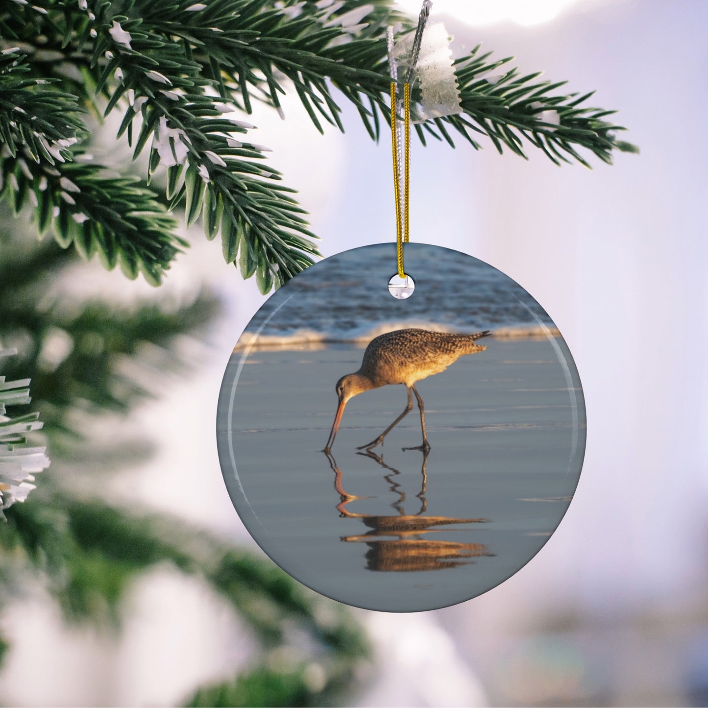 Ceramic holiday ornament featuring a photograph of a Long-billed Curlew mirrored in the wet sand of a shoreline under a soft golden sun, hanging on a Christmas tree branch.
