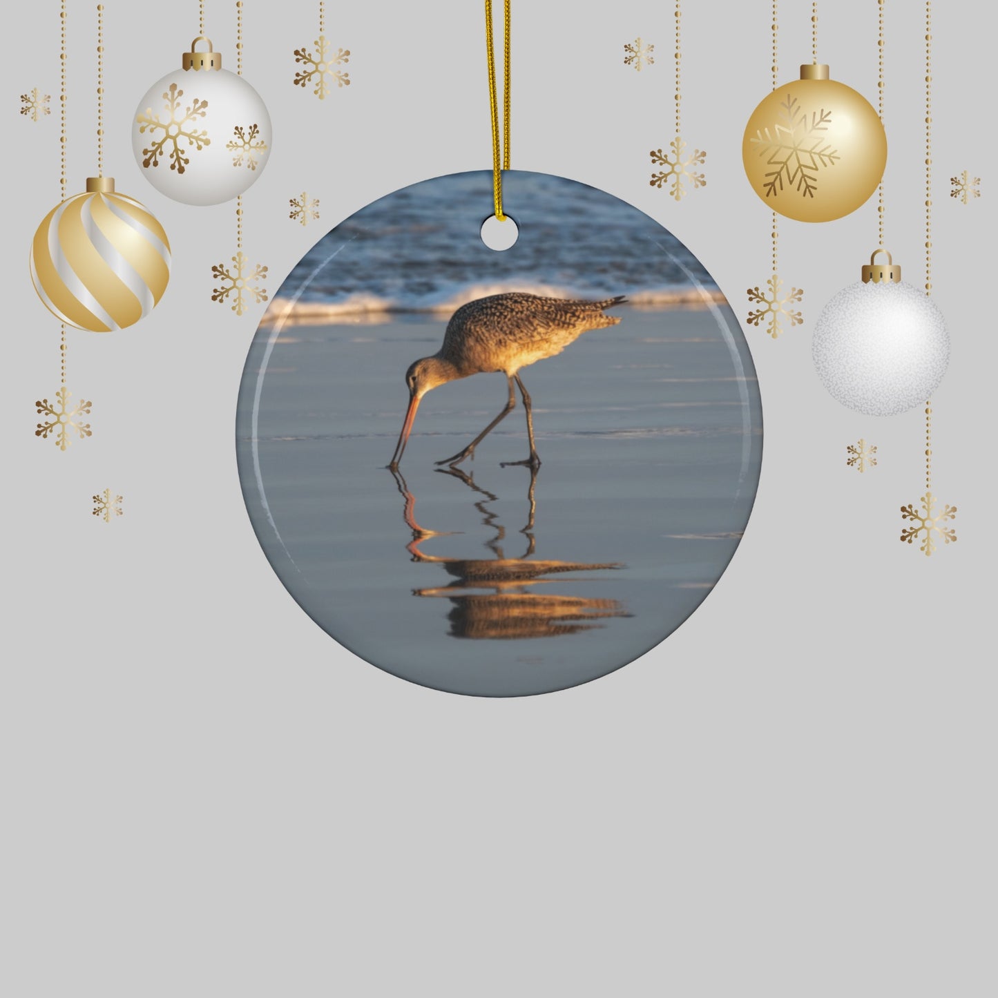 Ceramic holiday ornament featuring a photograph of a Long-billed Curlew mirrored in the wet sand of a shoreline under a soft golden sun and Christmas ornaments on a light gray background