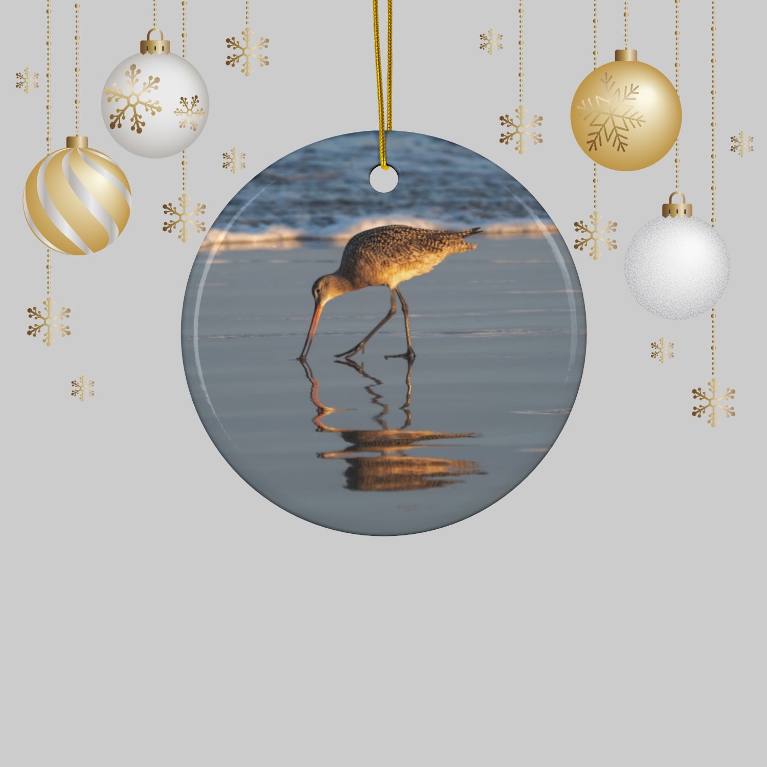 Ceramic holiday ornament featuring a photograph of a Long-billed Curlew mirrored in the wet sand of a shoreline under a soft golden sun and Christmas ornaments on a light gray background