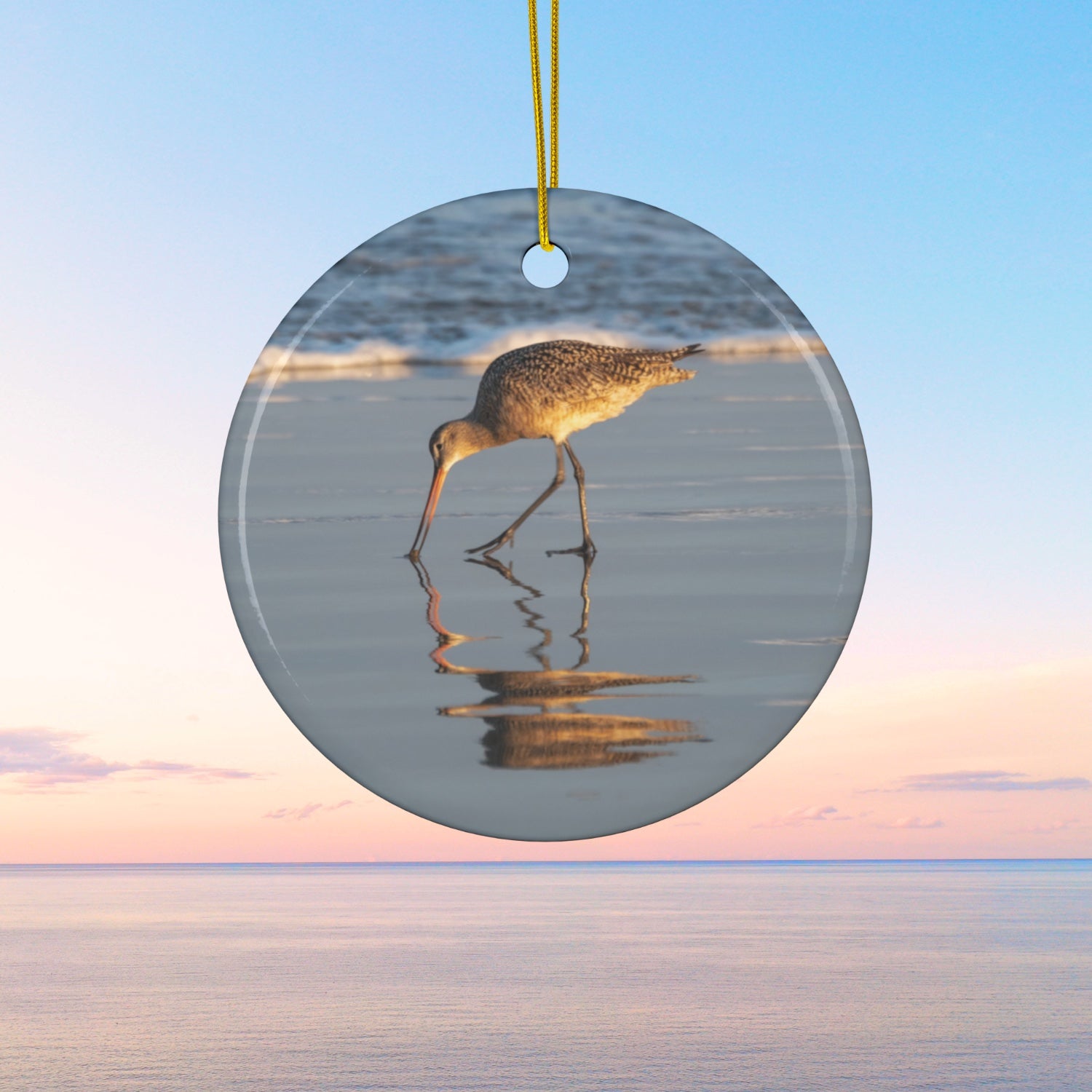 Ceramic holiday ornament featuring a photograph of a Long-billed Curlew mirrored in the wet sand of a shoreline under a soft golden sun