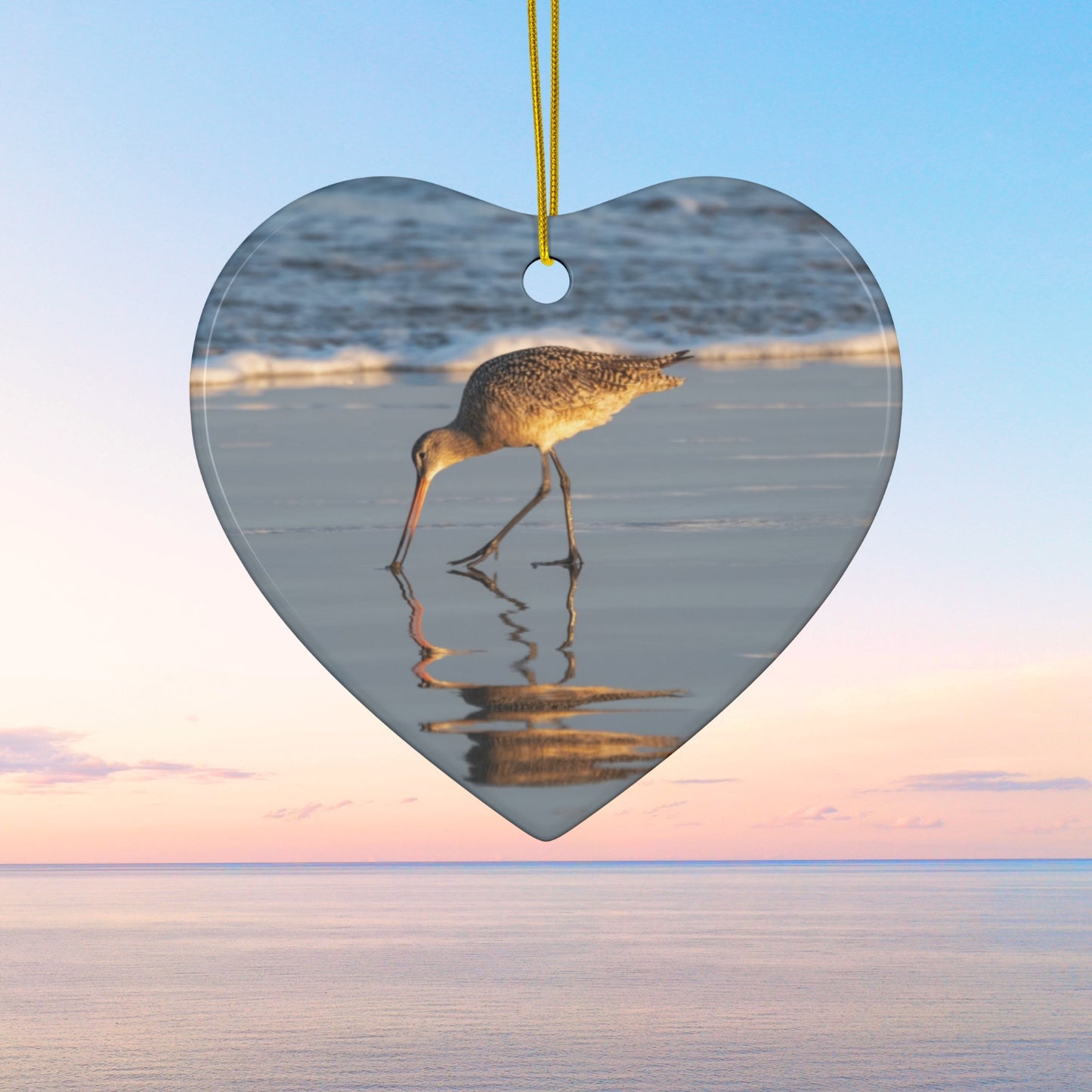 Heart-shaped ceramic holiday ornament featuring a photograph of a Long-billed Curlew mirrored in the wet sand of a shoreline under a soft golden sun