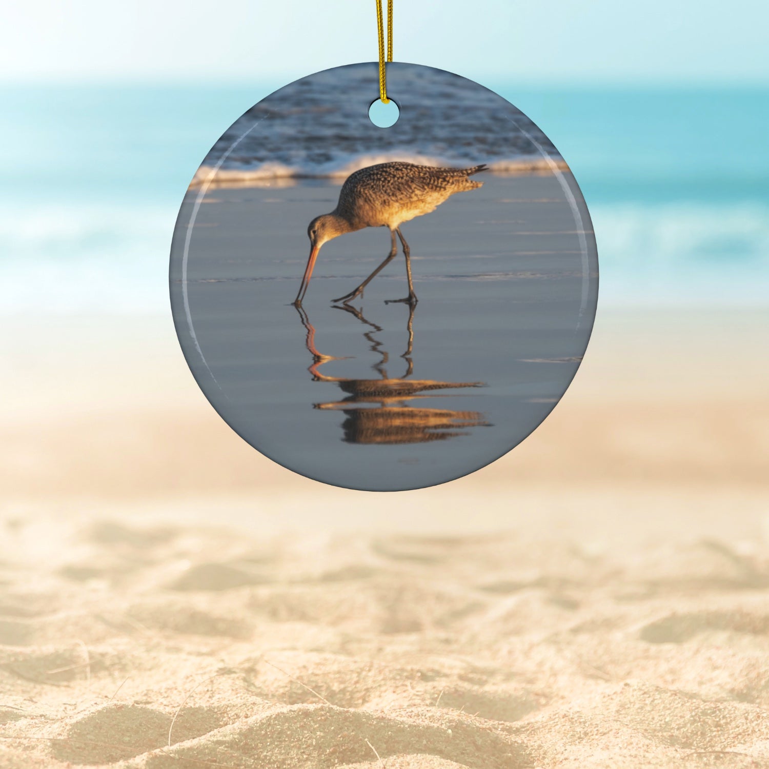 Ceramic holiday ornament featuring a photograph of a Long-billed Curlew mirrored in the wet sand of a shoreline under a soft golden sun
