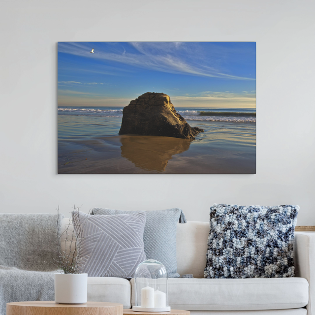 Canvas print of a rugged sea stack on a wet, reflective Malibu beach with a lone bird soaring in a clear blue sky over the Pacific Ocean on a wall above a sofa with decorative pillows.