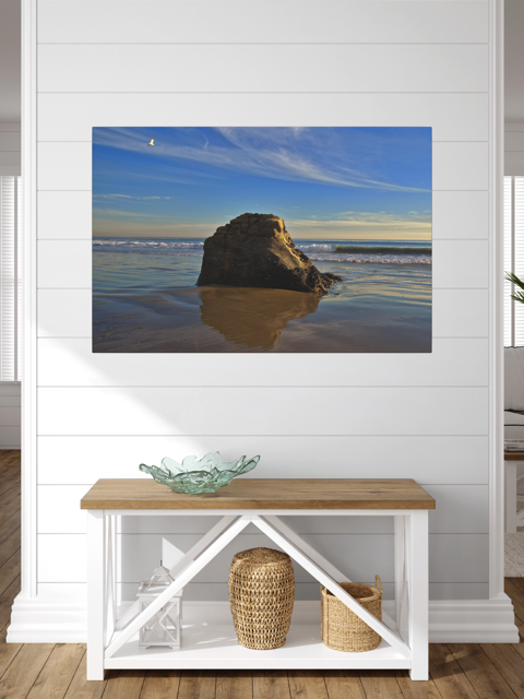 Canvas print of a rugged sea stack on a wet, reflective Malibu beach with a lone bird soaring in a clear blue sky over the Pacific Ocean on a wall above a wooden bench with storage baskets.
