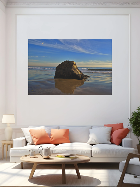 Living room with a lcanvas print of a rugged sea stack on a wet, reflective Malibu beach with a lone bird soaring in a clear blue sky over the Pacific Ocean on the wall above a white sofa with colorful cushions.