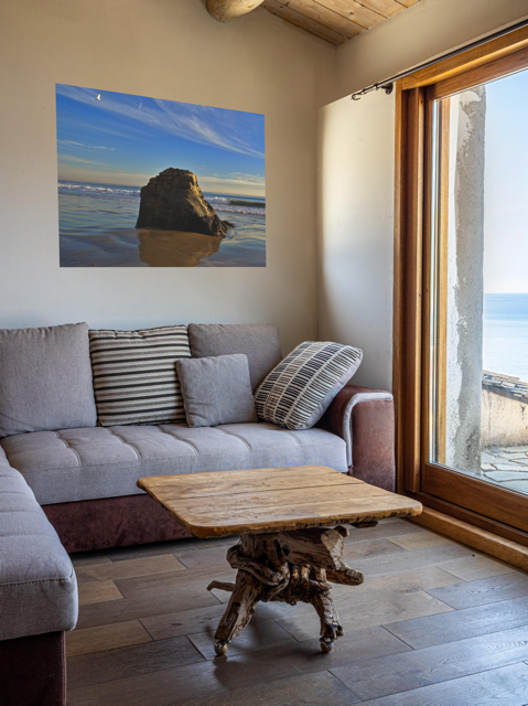 Living room with a gray sofa, wooden coffee table, and a canvas print of a rugged sea stack on a wet, reflective Malibu beach with a lone bird soaring in a clear blue sky over the Pacific Ocean.