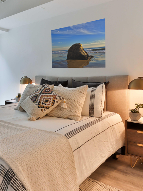 Bedroom with a bed featuring patterned pillows, a headboard, and a canvas print of a rugged sea stack on a wet, reflective Malibu beach with a lone bird soaring in a clear blue sky over the Pacific Ocean.