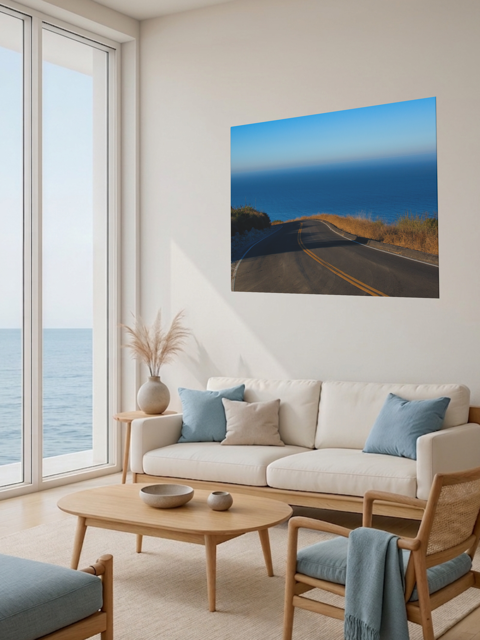Living room with a large window view of the ocean, a beige sofa with blue cushions, and a canvas print of a winding mountain road above the Pacific Ocean in Malibu California.