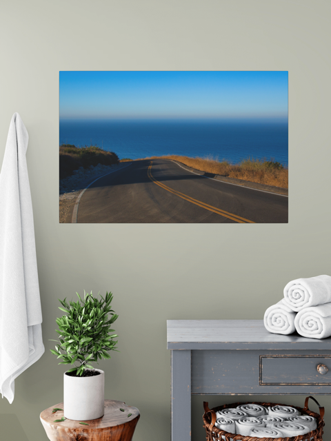 Canvas print of a winding mountain road above the Pacific Ocean in Malibu California on a wall above a table with a plant and folded towels.