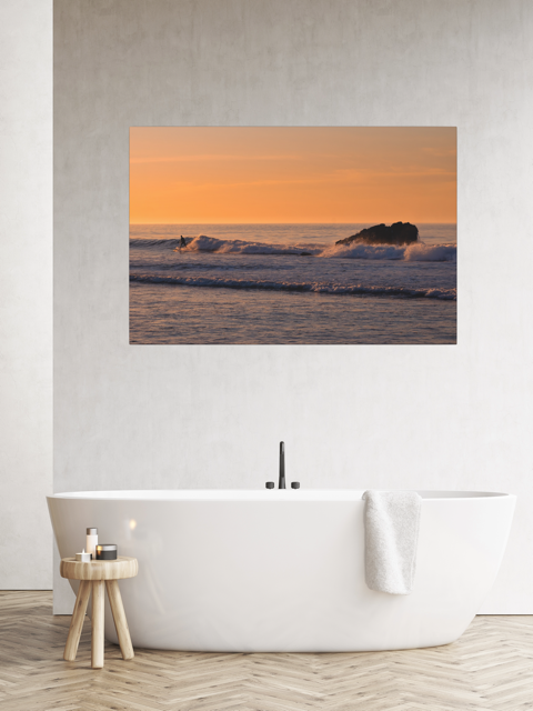 Bathroom with a canvas print featuring a surfer riding a wave at sunrise, a white bathtub, and a small wooden stool.