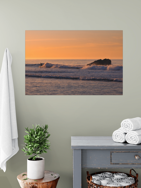 Canvas print featuring a surfer riding a wave at sunrise on a wall above a table with towels and a plant.
