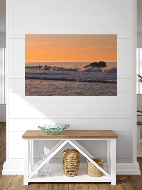 Canvas print featuring a surfer riding a wave at sunrise on a white wall above a wooden bench with storage baskets.