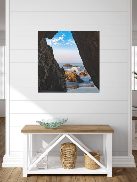 A canvas print of a view from inside a dark Malibu sea cave looking out at crashing ocean waves and rocks under a blue sky on a white wall above a wooden bench.