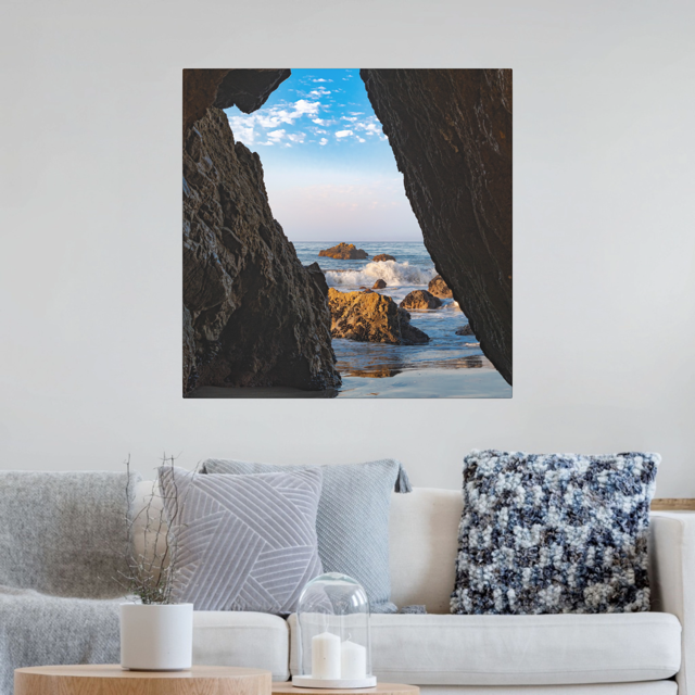 Living room with a couch and decorative pillows, featuring a canvas print of a view from inside a dark Malibu sea cave looking out at crashing ocean waves and rocks under a blue sky.