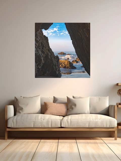 Sofa with decorative pillows in a room with a canvas print of a view from inside a dark Malibu sea cave looking out at crashing ocean waves and rocks under a blue sky.