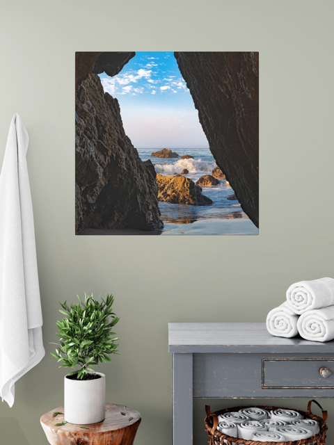 A canvas print of a view from inside a dark Malibu sea cave looking out at crashing ocean waves and rocks under a blue sky as wall art in a room with a plant and towels.