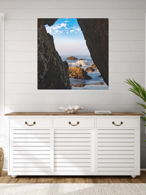 White cabinet with wooden top against a wall with a canvas print of a view from inside a dark Malibu sea cave looking out at crashing ocean waves and rocks under a blue sky.