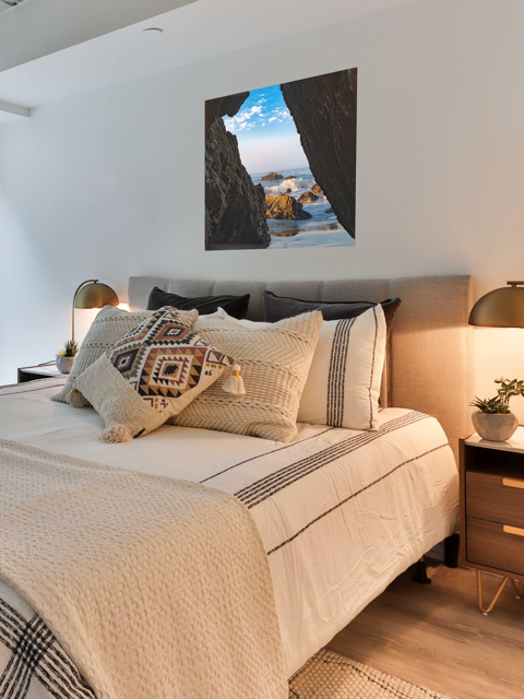 Neatly made bed with decorative pillows in a bedroom setting with a canvas print of a view from inside a dark Malibu sea cave looking out at crashing ocean waves and rocks under a blue sky.