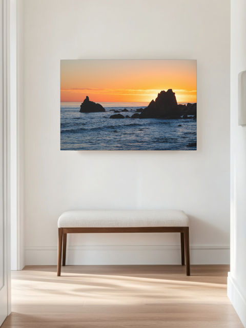 Canvas print of an ocean sunset with rocks, displayed on a wall above a bench.