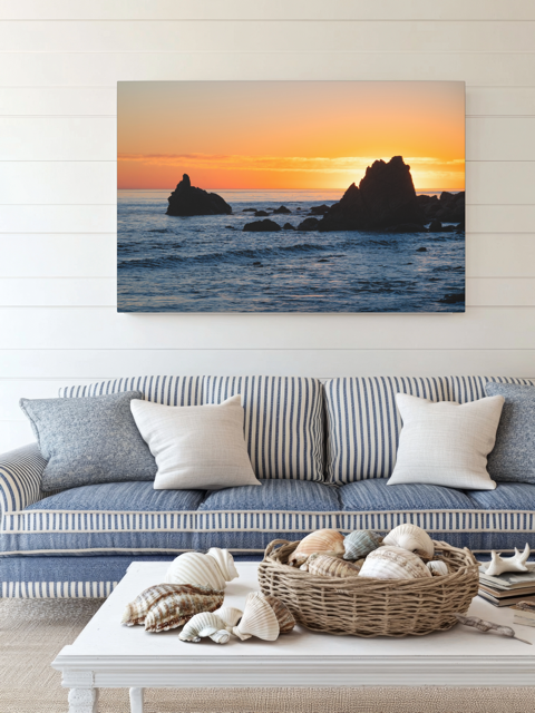 Canvas print of an ocean sunset with rocks displayed over a blue sofa.