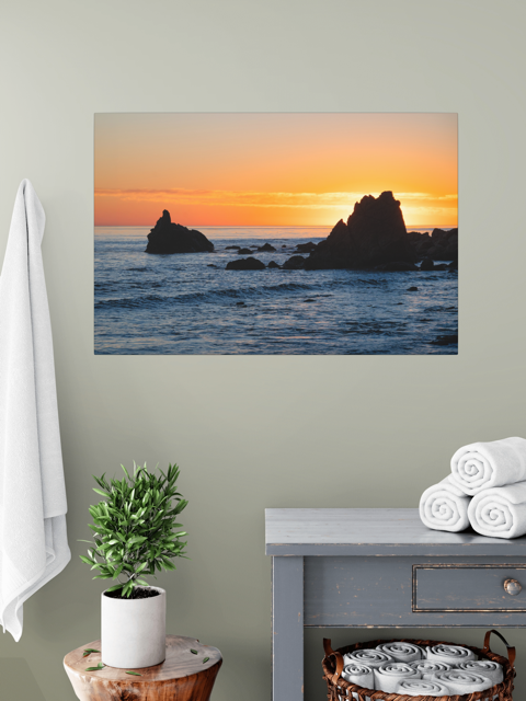 Canvas print of an ocean sunset with rocks on wall above a plant and cabinet.