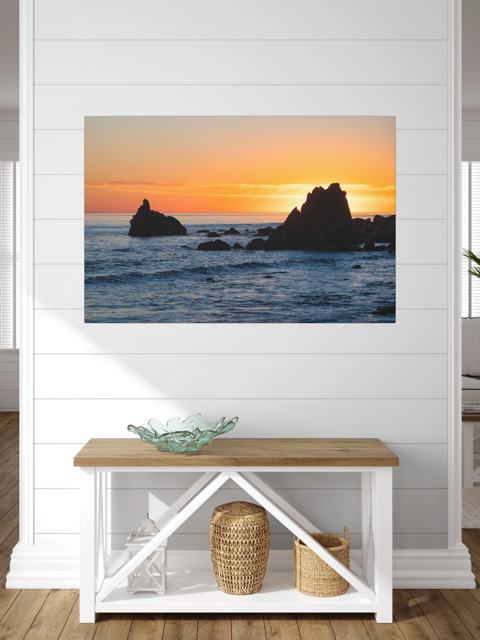 Canvas print of an ocean sunset with rocks on a white wall above a wooden bench.