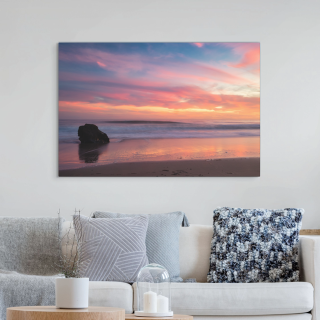 Canvas print of a solitary rock sea stack on a Malibu beach at sunset with a vibrant pink, orange, and purple sky reflected in the wet sand hanging on a wall above a sofa.