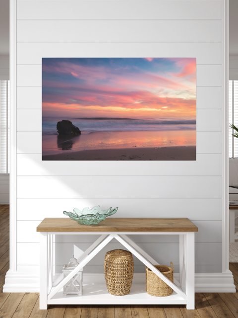 Canvas print of a solitary rock sea stack on a Malibu beach at sunset with a vibrant pink, orange, and purple sky reflected in the wet sand on a wall above a wooden bench with storage baskets.