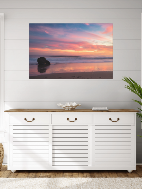 White cabinet with wooden top against a wall with a canvas print of a solitary rock sea stack on a Malibu beach at sunset with a vibrant pink, orange, and purple sky reflected in the wet sand.