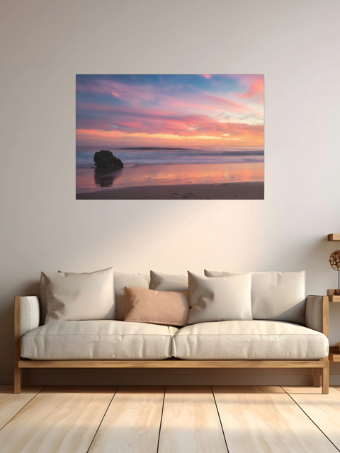 Canvas print of a solitary rock sea stack on a Malibu beach at sunset with a vibrant pink, orange, and purple sky reflected in the wet sand on a wall above a sofa with cushions.