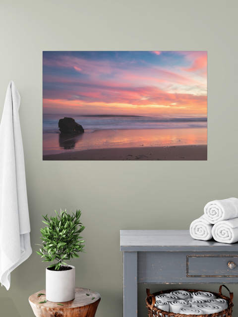 Canvas print of a solitary rock sea stack on a Malibu beach at sunset with a vibrant pink, orange, and purple sky reflected in the wet sand on a wall above a table with a plant and folded towels.