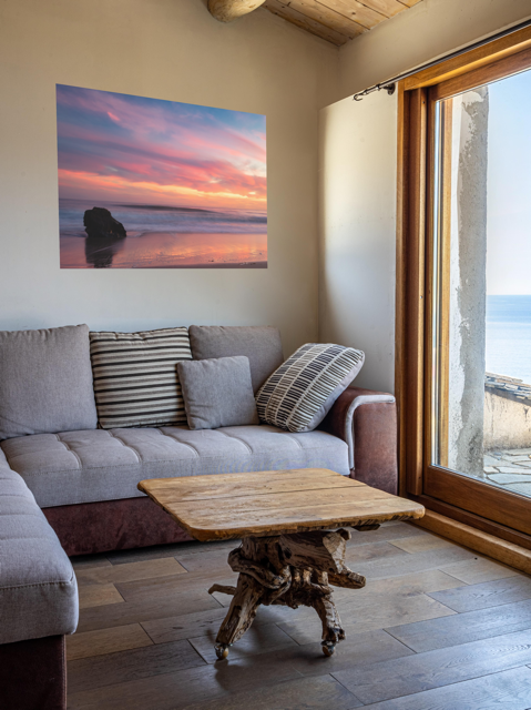 Living room with a gray sofa, wooden coffee table, and a canvas print of a solitary rock sea stack on a Malibu beach at sunset with a vibrant pink, orange, and purple sky reflected in the wet sand.