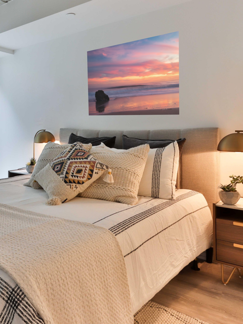 Bedroom with a bed featuring decorative pillows and a canvas print of a solitary rock sea stack on a Malibu beach at sunset with a vibrant pink, orange, and purple sky reflected in the wet sand on the wall.