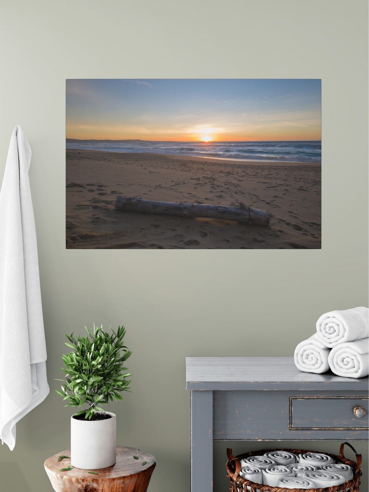 Canvas print of a Monterey California beach sunset with large driftwood log on sand on a wall above a table with a plant and folded towels.