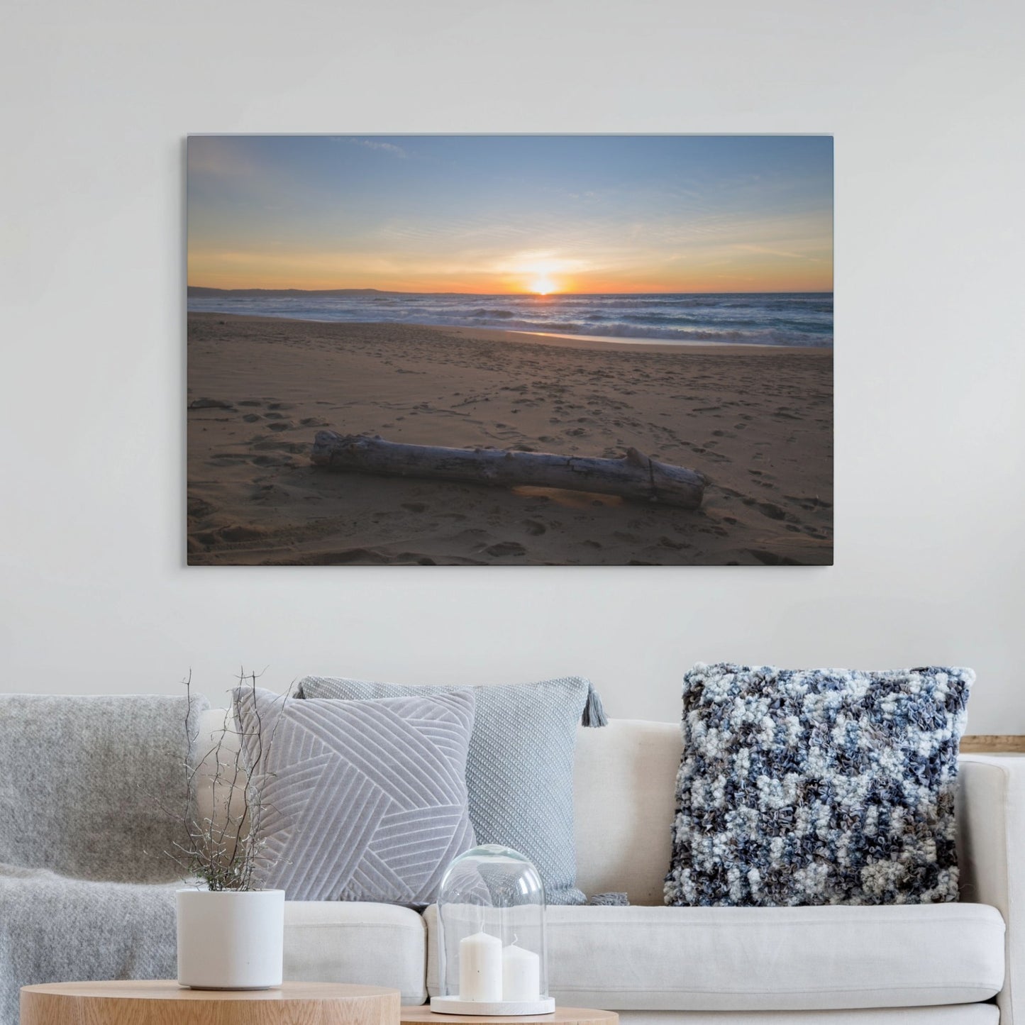 Canvas print of a Monterey California beach sunset with large driftwood log on sand on a wall above a sofa with decorative pillows.