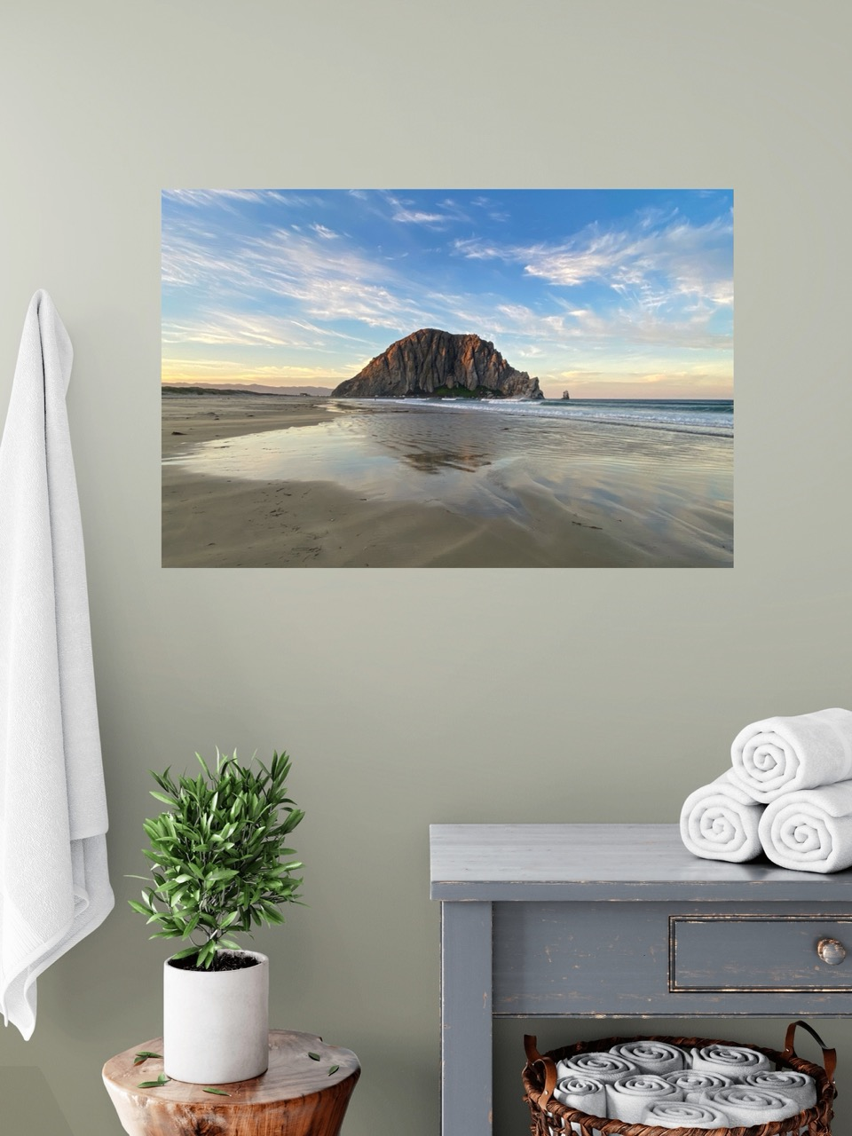 Canvas print of Morro Rock from the beach in Morro Bay, California with reflections in the sand and a colorful sky on a wall above a dresser with towels and a plant