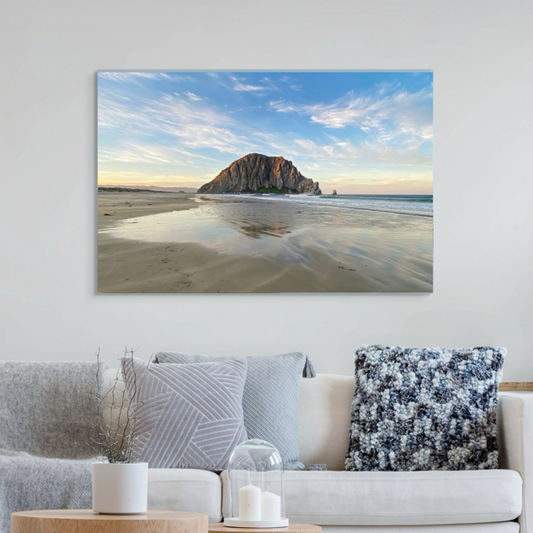 Large canvas print of Morro Rock from the beach in Morro Bay, California with reflections in the sand and a colorful sky on a wall above a sofa with decorative pillows.