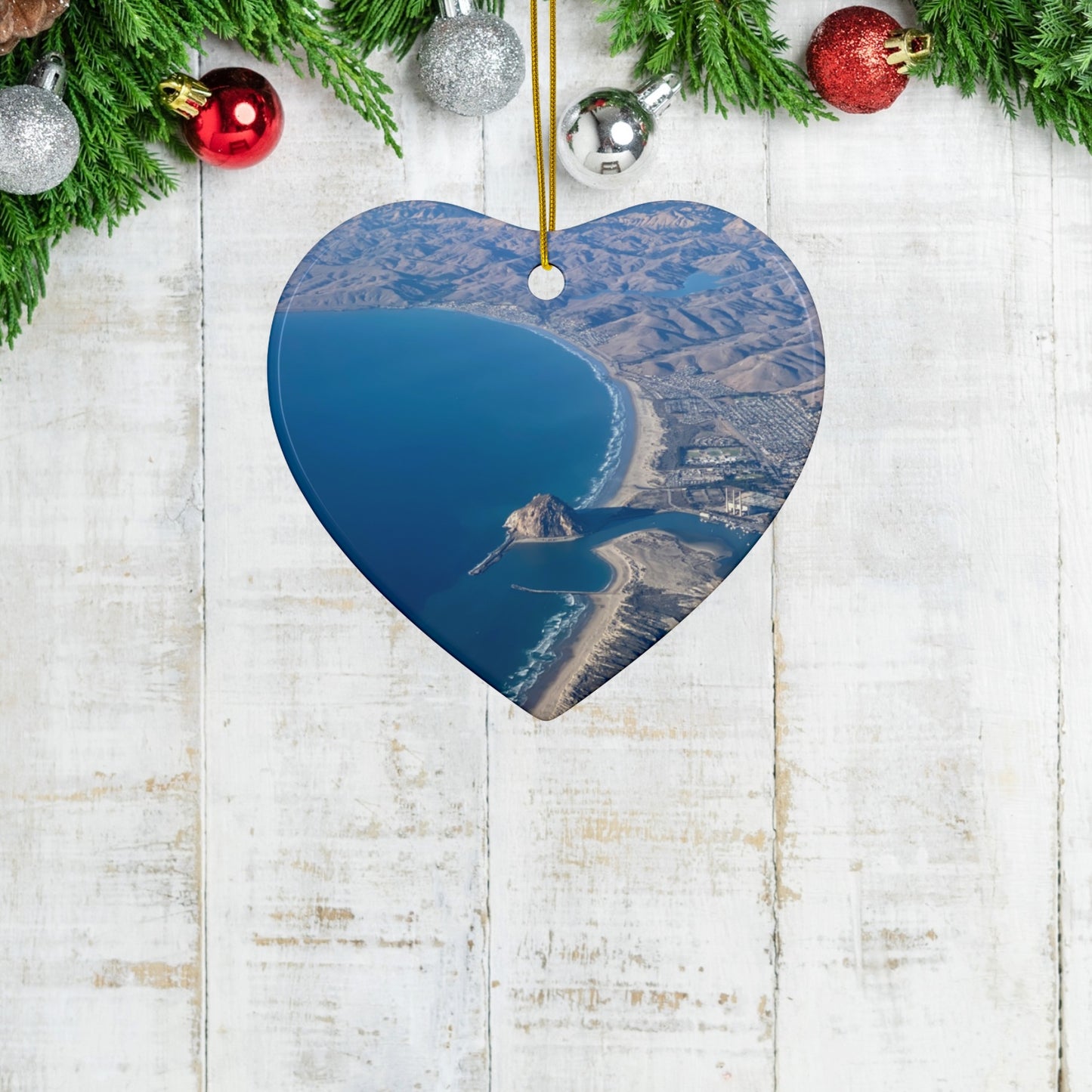 Heart-shaped ceramic holiday ornament featuring an aerial photograph of Morro Rock and the coastline of Morro Bay, California, with vibrant blue water and golden hills on a wooden surface.