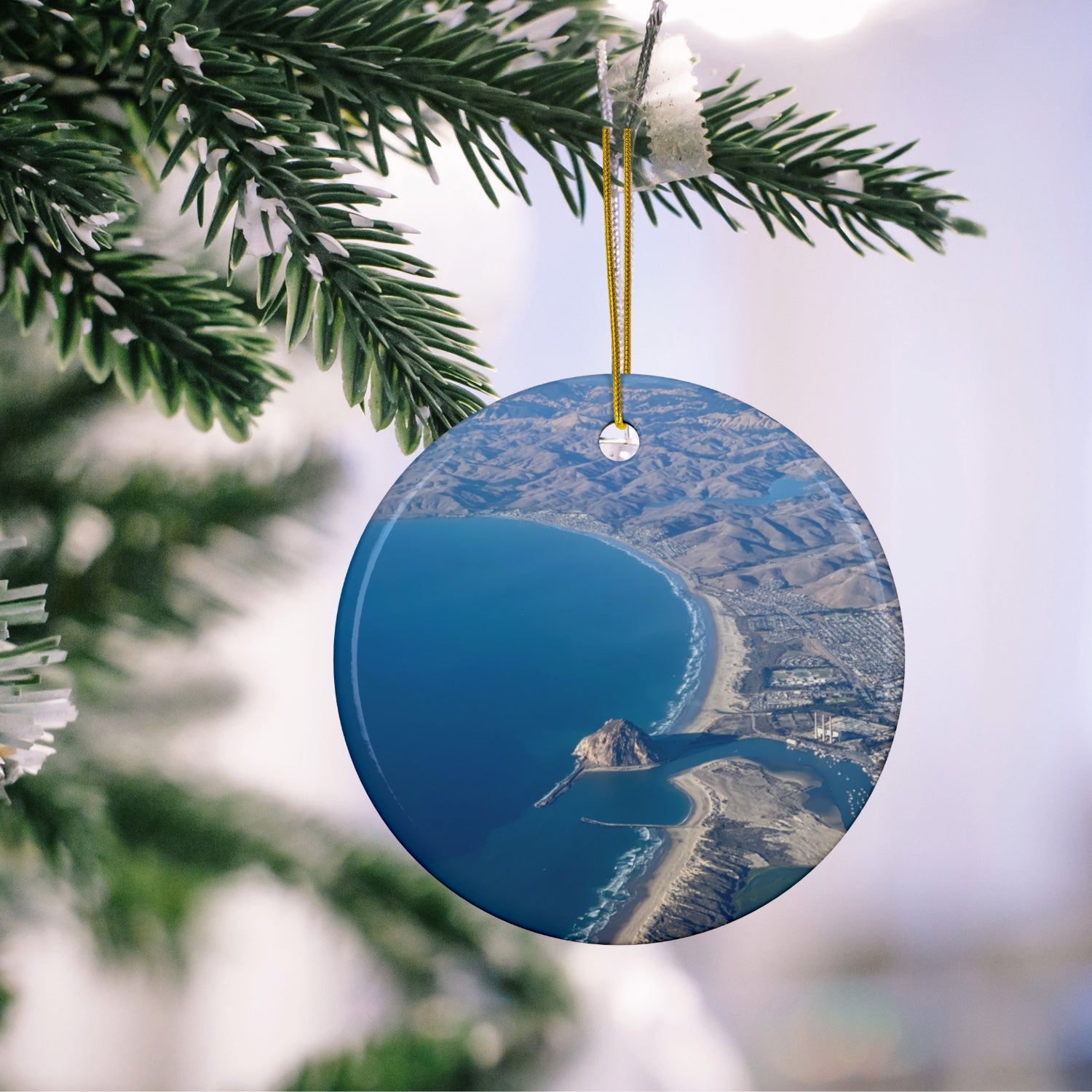 Ceramic holiday ornament featuring an aerial photograph of Morro Rock and the coastline of Morro Bay, California, with vibrant blue water and golden hills, hanging on a Christmas tree.