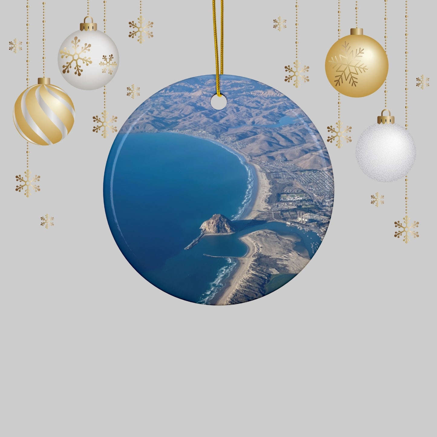 Ceramic holiday ornament featuring an aerial photograph of Morro Rock and the coastline of Morro Bay, California, with vibrant blue water and golden hills with Christmas ornaments on a light gray background.
